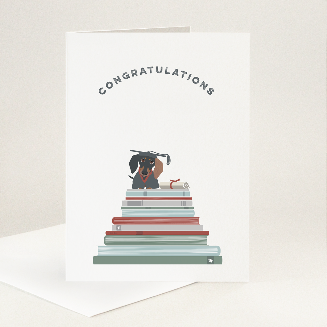 A little sausage dog wears a mortar board and gown and is sat on a pile of books with a scroll next to them all under the word congratulations to celebrate their graduation. 